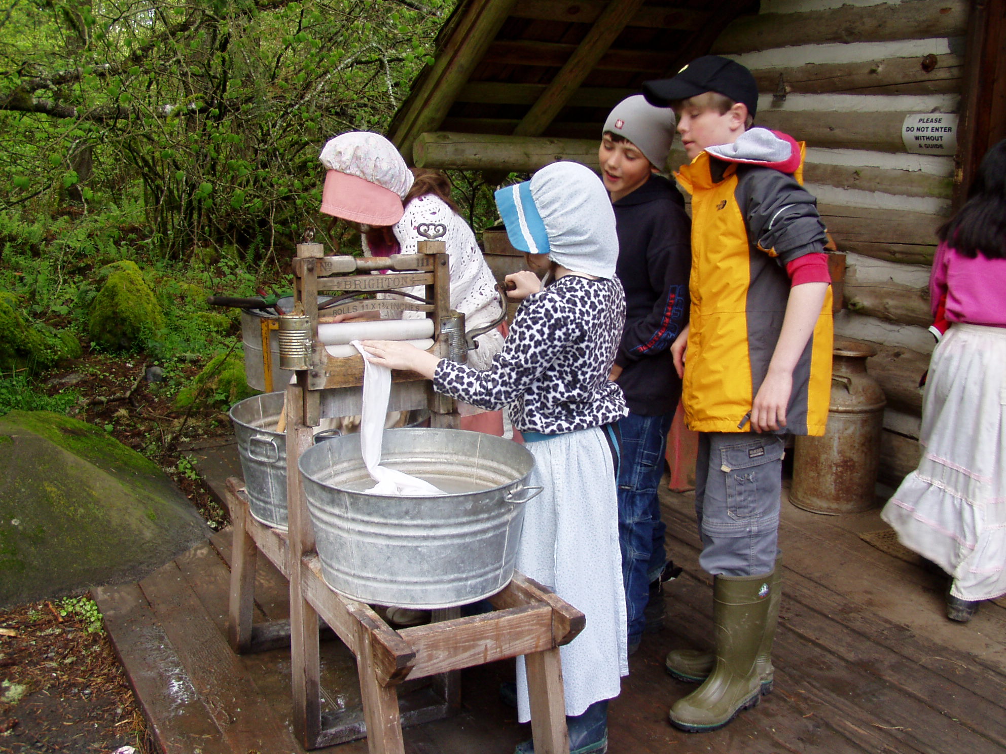 two students in sun bonnets using washtubs outside a cabin, while two others watch
